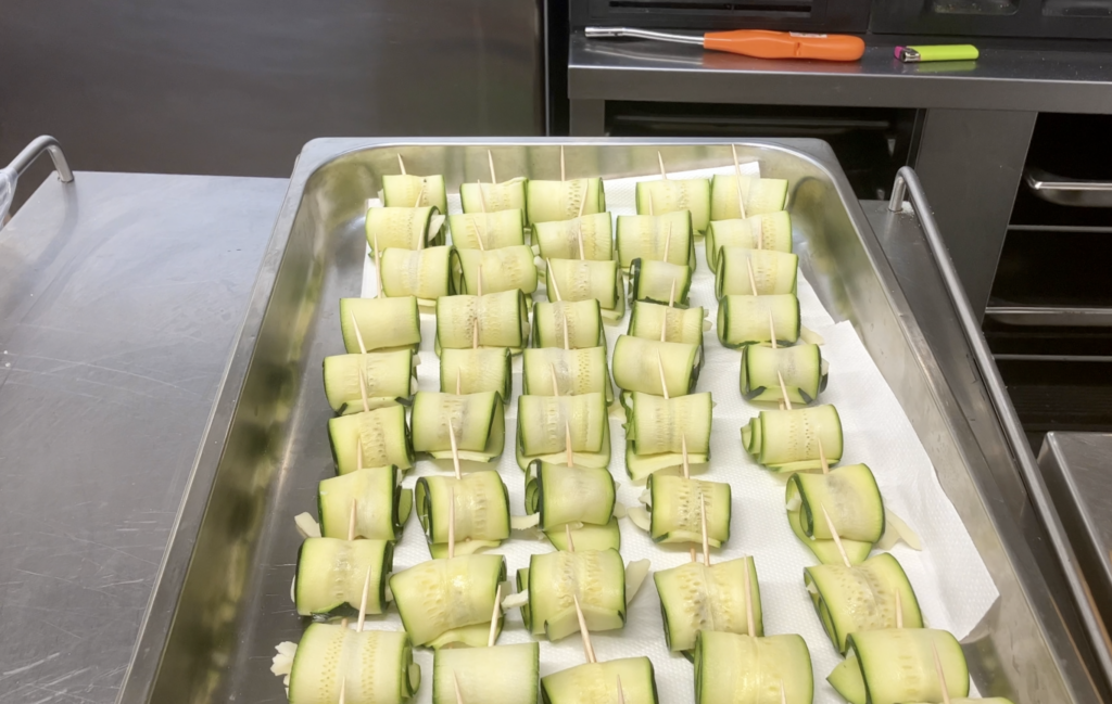 Forty-five thin cross-sections of zucchini, wrapped into cylinders and skewered with toothpicks, sit in a hotel pan on a metal countertop.